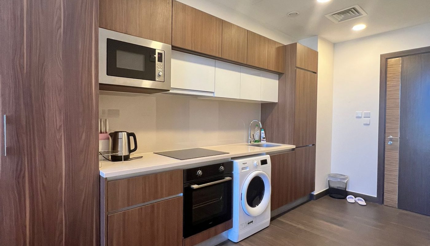 Modern kitchen with wooden cabinets, built-in oven, electric stove, microwave, kettle, washing machine, and sink; minimal accessories, brown flooring, and closed doors visible.