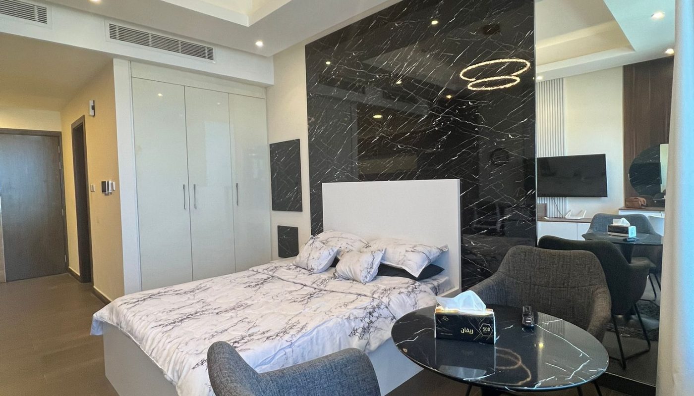 Modern bedroom with marble accent wall, circular ceiling lights, a bed with white bedding, a round glass table, two gray chairs, and a wardrobe with mirrored doors.