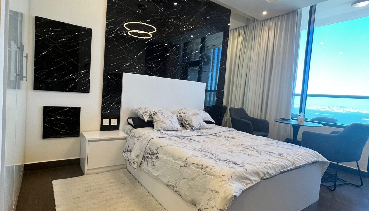 Modern bedroom with a large bed, marble accent wall, circular ceiling light, seating area, floor-to-ceiling window, and dark wood flooring.