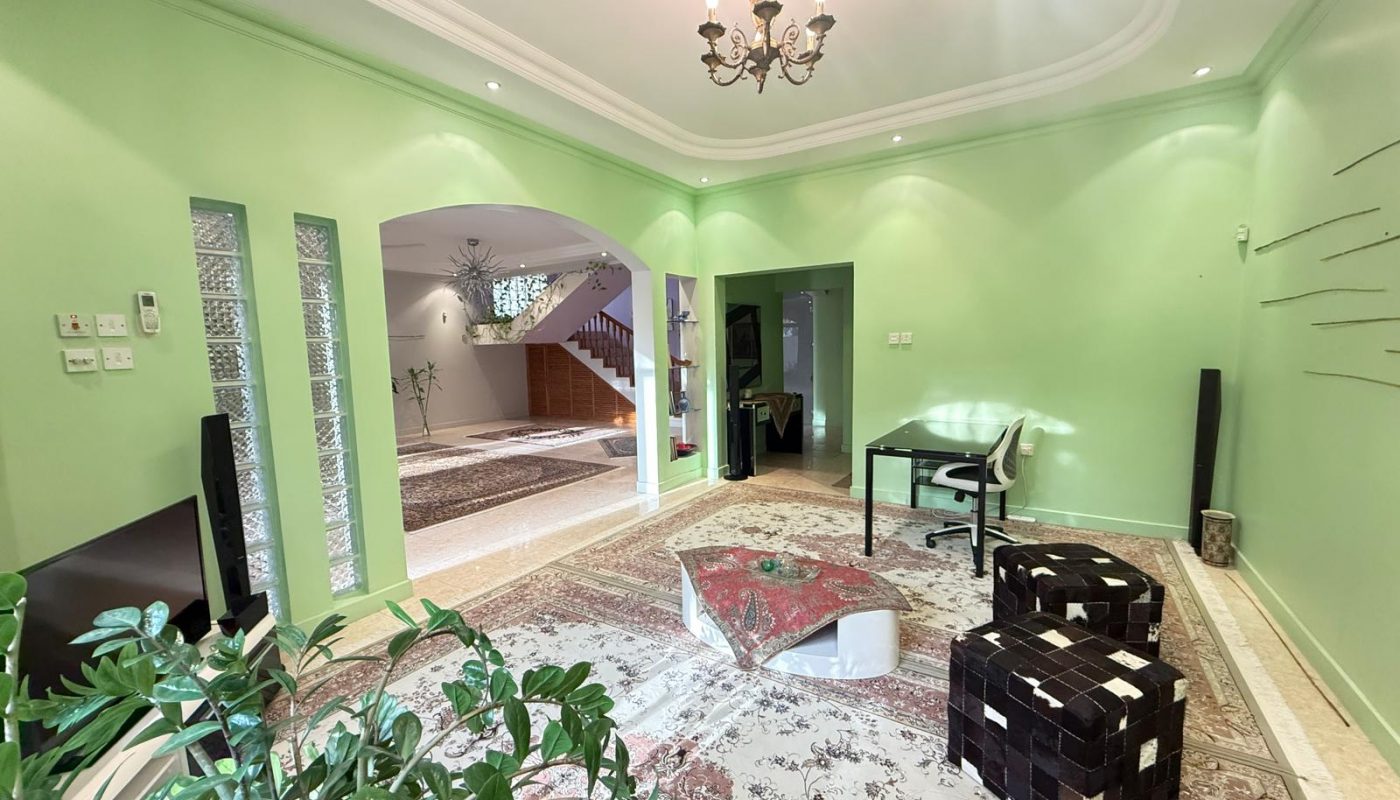 A spacious living room with light green walls, patterned rugs, a desk, two woven chairs, a low table, a TV, houseplants, and an open archway leading to a staircase.