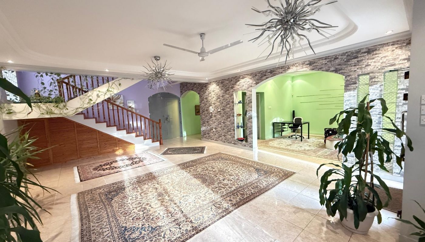 Spacious, well-lit room with tiled floor, patterned rugs, green accent walls, indoor plants, modern light fixtures, and a wooden staircase with railing.