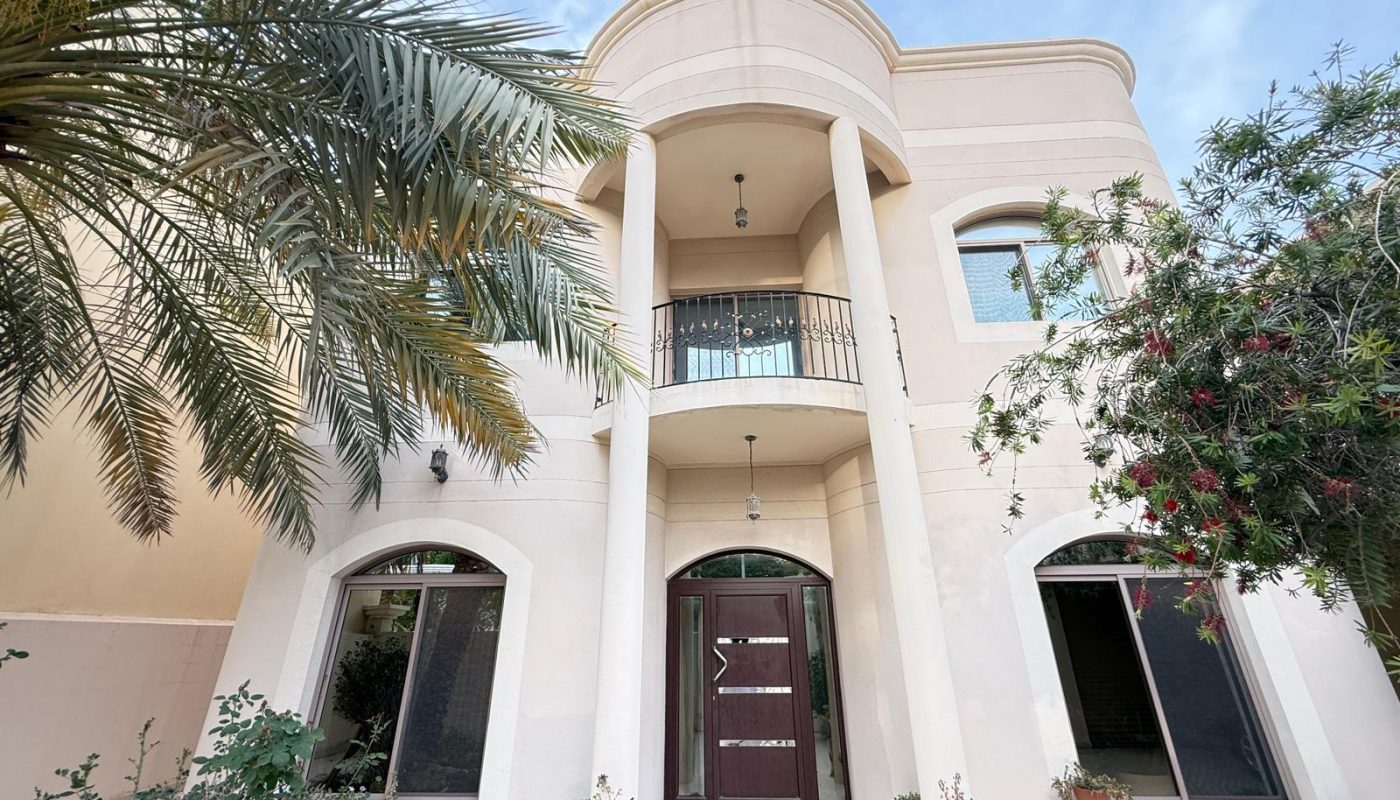 Two-story beige house with arched windows, a central balcony, double front doors, columns, and surrounding plants under a blue sky.