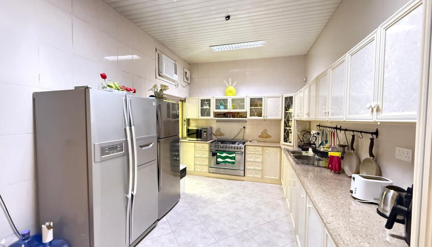 A modern kitchen with white cabinets, stainless steel appliances, a double-door refrigerator, oven, microwave, and countertop appliances, with a water dispenser and tiled flooring.