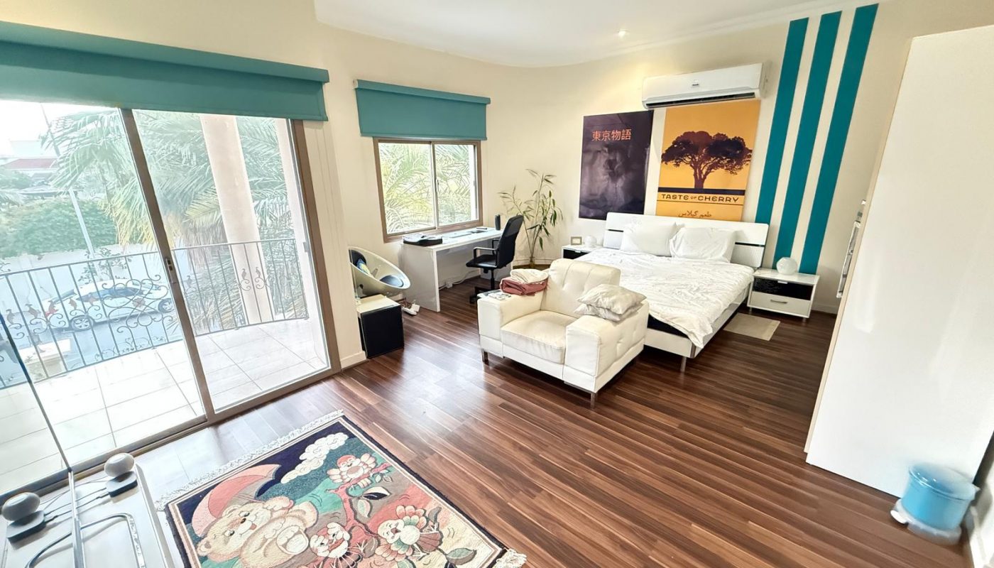Spacious bedroom with wood flooring, double bed, white sofa, desk, balcony doors, large windows, and colorful wall art.