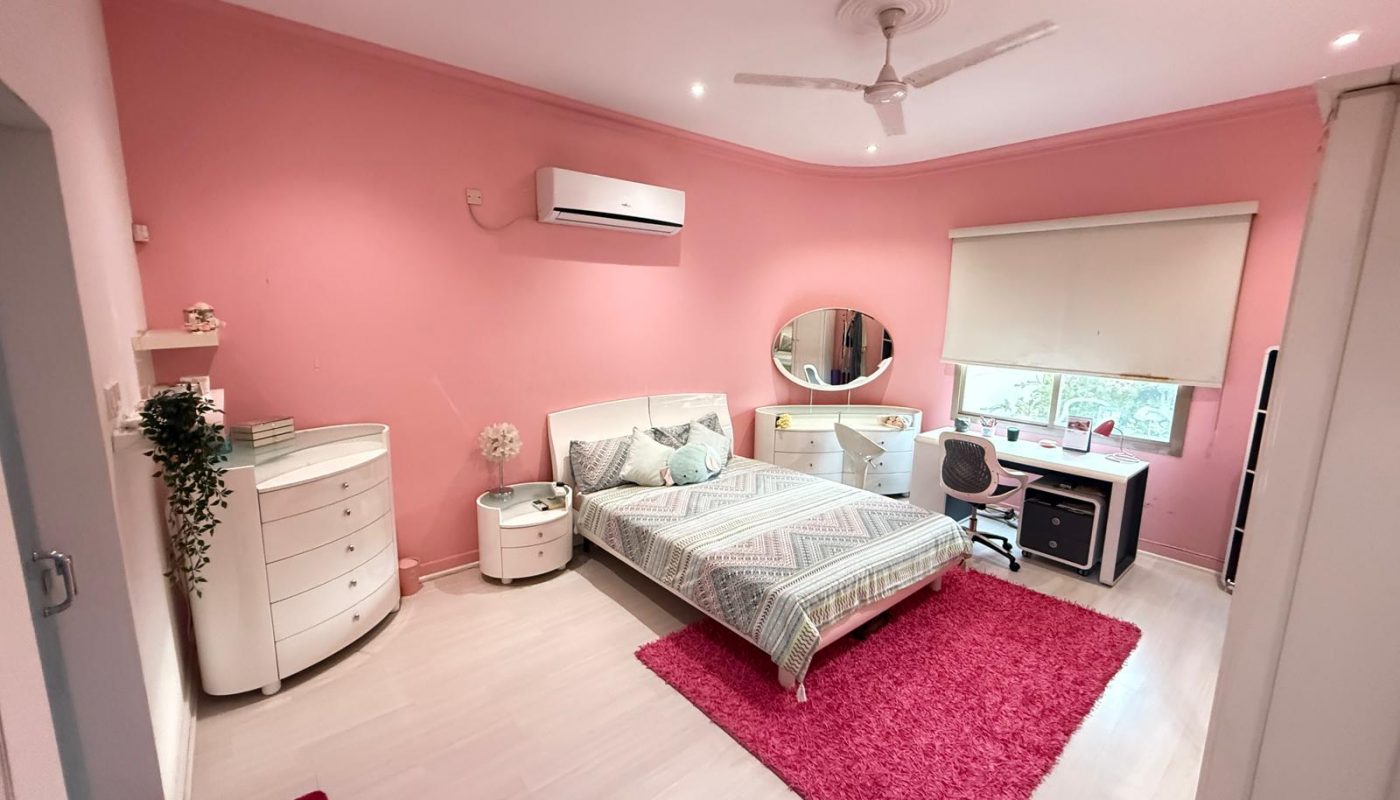 A bedroom with pink walls, a single bed, white furniture, a pink rug, a desk near the window, and a ceiling fan.