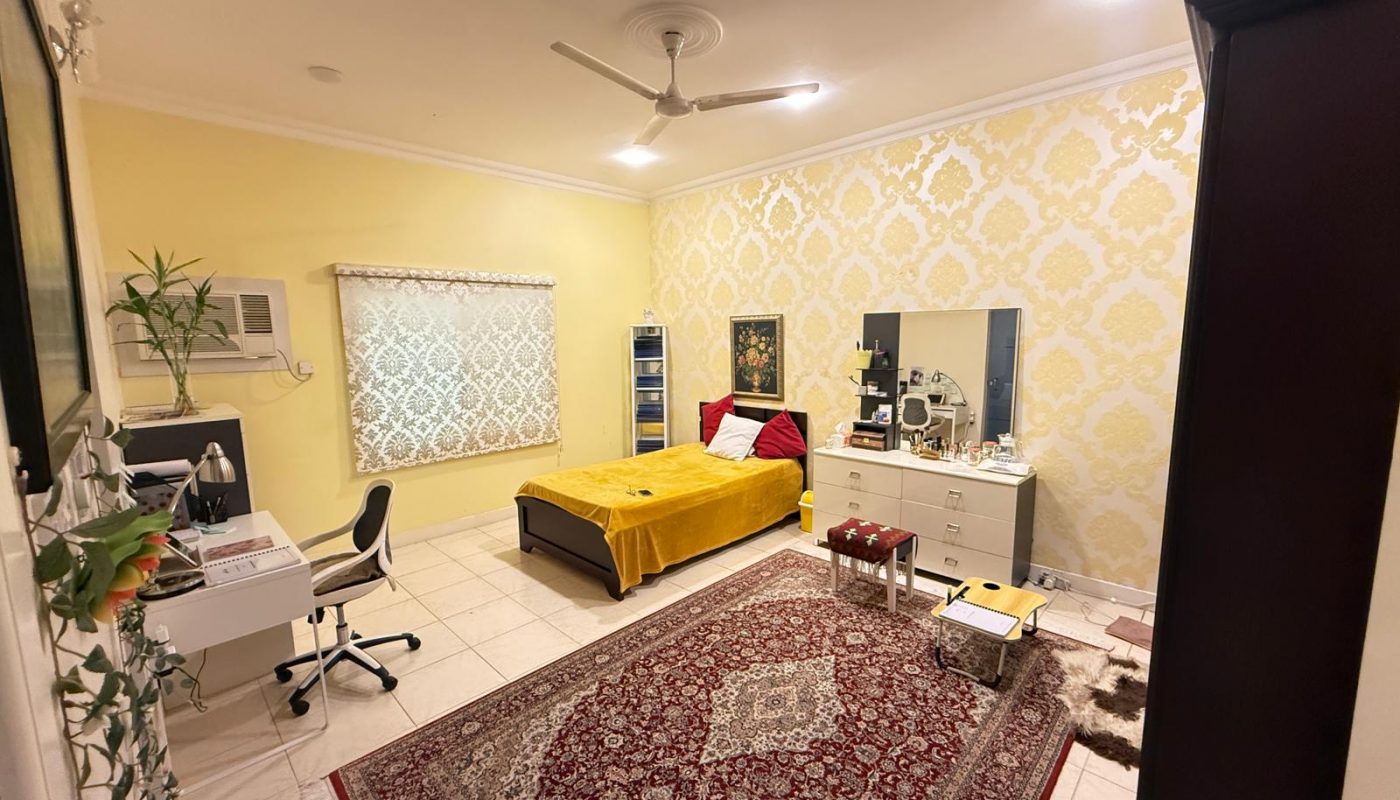 A well-lit bedroom with a bed, desk, chair, dresser, wall art, and a patterned rug on a tiled floor. The walls are yellow with ornate wallpaper on one side.