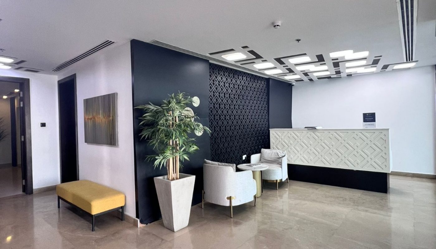 Modern office reception area with a patterned wall, front desk, two white chairs, potted plant, and a yellow bench on a polished marble floor.