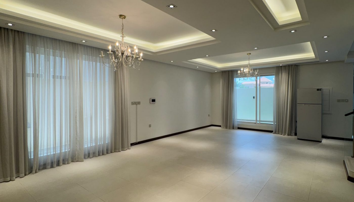Spacious empty living room with large tile flooring, ceiling lights, chandeliers, floor-to-ceiling curtains, and a refrigerator near sliding glass doors.