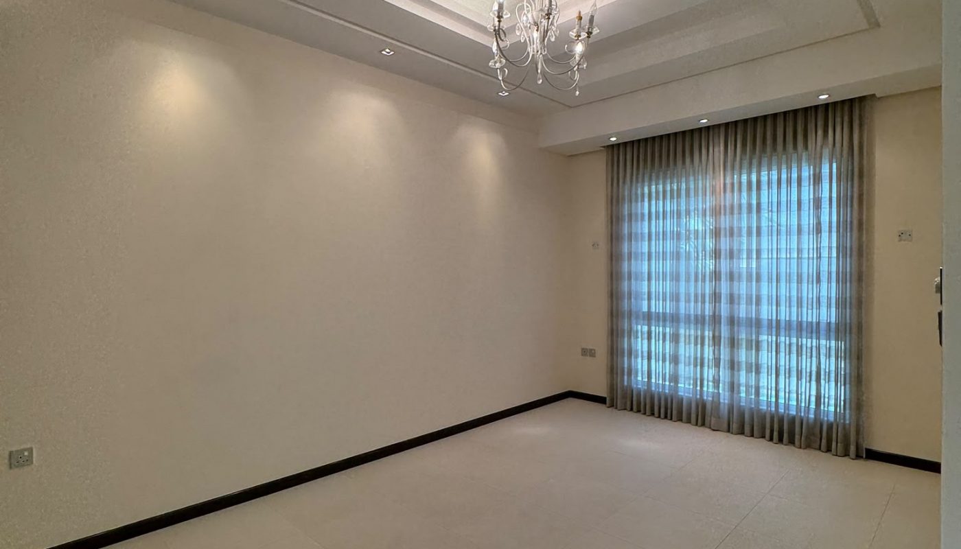 Empty room with cream-colored walls and tiled floor, a ceiling chandelier, recessed lights, and a large window covered with sheer curtains letting in natural light.