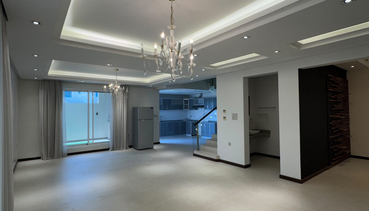 Spacious, modern living room with tiled floor, recessed and chandelier lighting, large windows with sheer curtains, and a staircase leading to the upper floor.