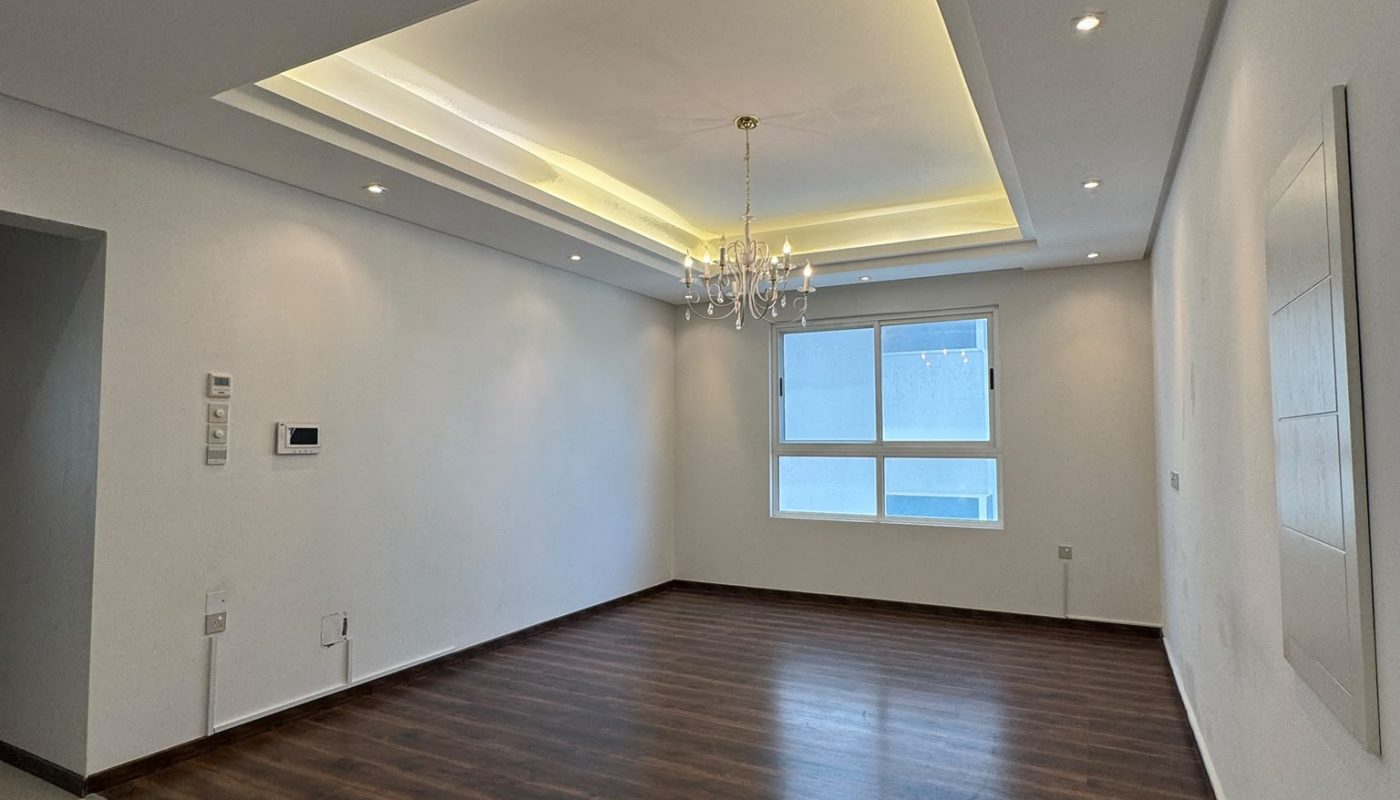 A modern, empty room with wooden flooring, white walls, recessed ceiling lights, a chandelier, and a large window with daylight coming through.