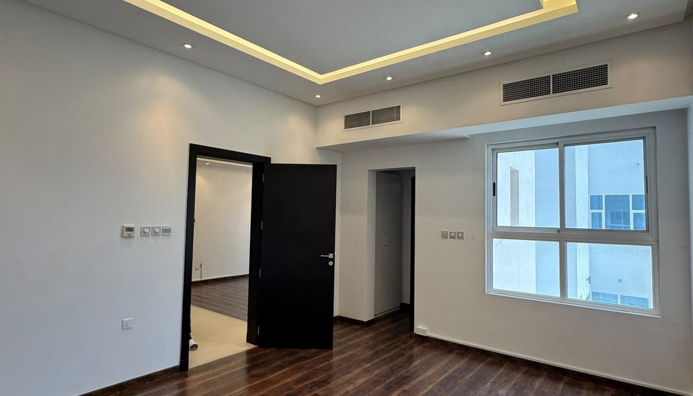 A modern, empty room with wood flooring, white walls, a large window, recessed ceiling lights, and an open black door leading to another space.