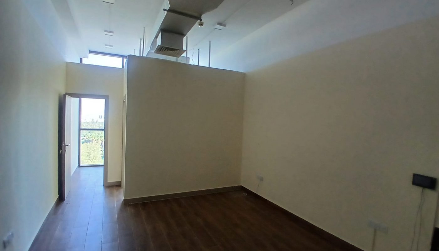Empty room with wooden floor, cream-colored walls, high ceiling, visible ventilation ducts, and an open door leading to a balcony with a view of greenery.