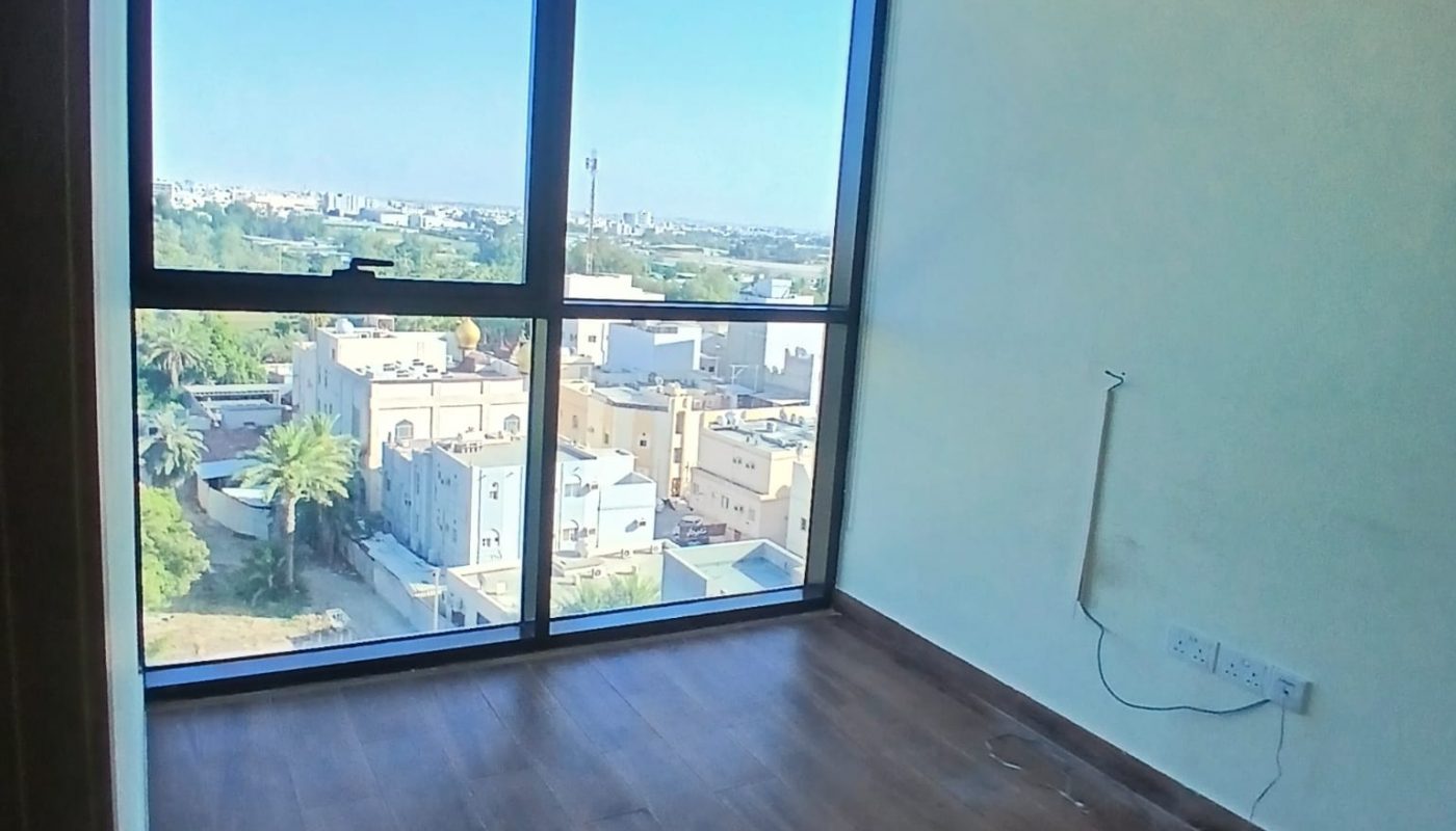 Empty room with wooden flooring, large floor-to-ceiling windows, a view of buildings and trees outside, and exposed cables on the wall.