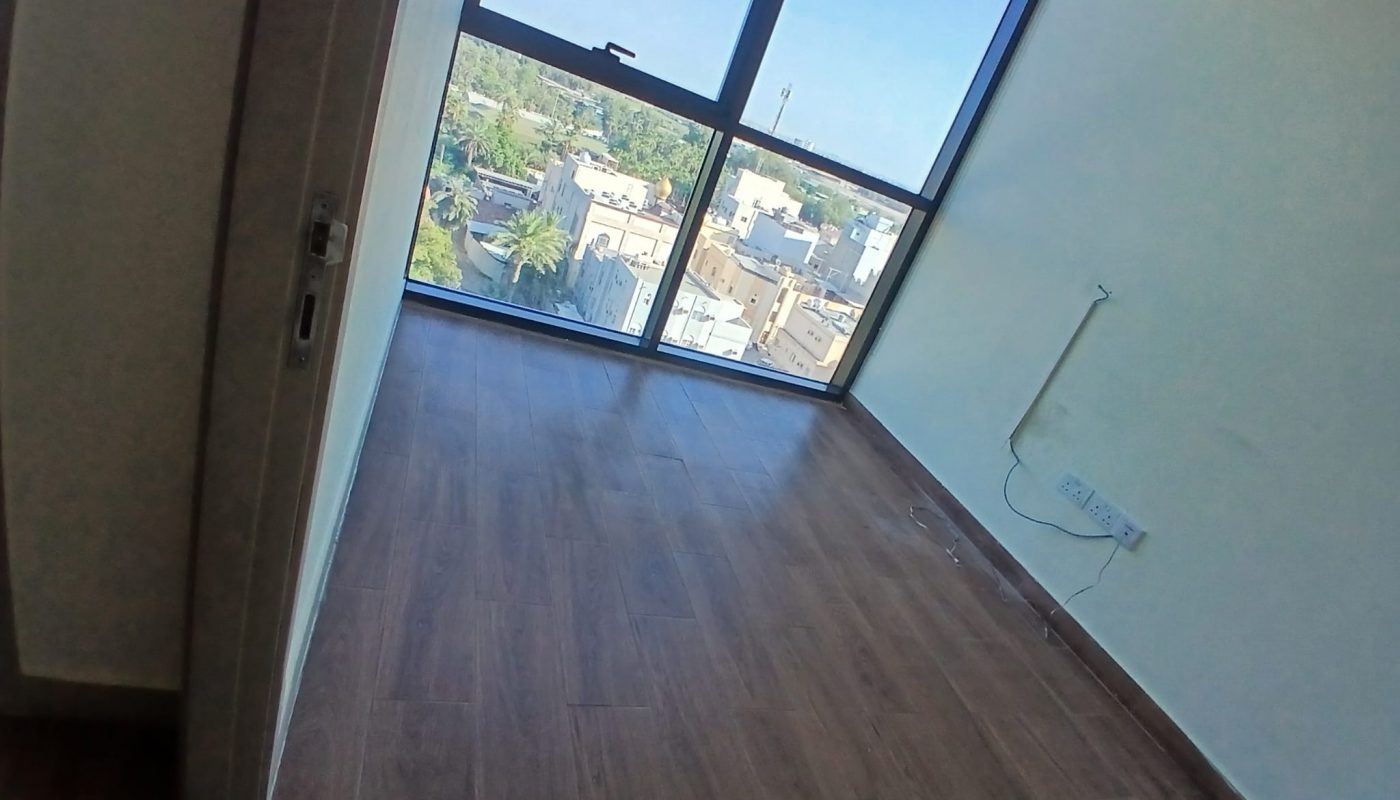 Empty room with wooden flooring, a large floor-to-ceiling window showing a cityscape, and a wall with electrical outlets and cables.