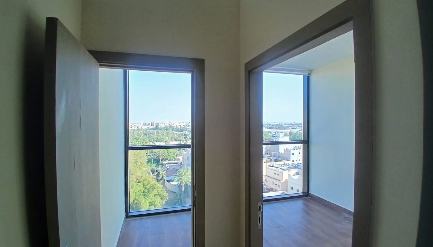 Two adjacent rooms with large windows and wooden floors are visible from an open doorway, overlooking a cityscape with greenery outside.