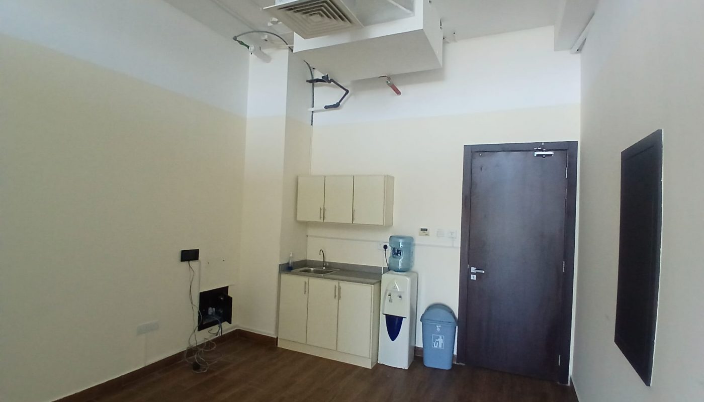 Small break room with a sink, cabinets, water dispenser, trash bin, and a closed dark door; several exposed pipes and vents visible on the ceiling.