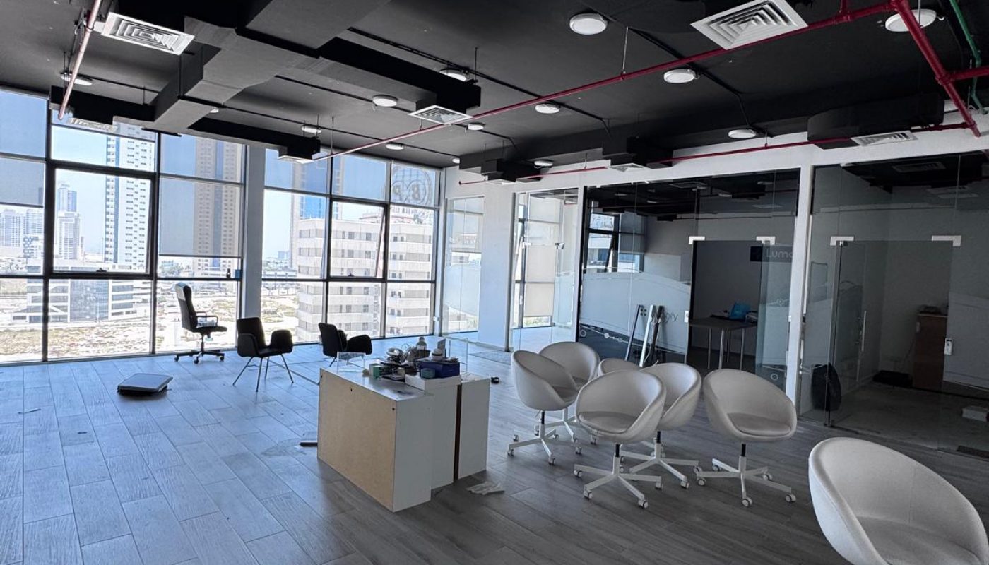 Modern office space with empty swivel chairs, a central desk, and large windows overlooking city buildings during daytime. Some items are scattered on the floor.