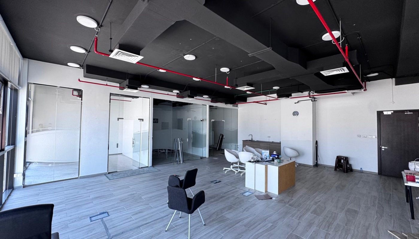 Modern office space with glass-walled meeting rooms, an uncluttered central area, scattered chairs, a desk, and exposed ceiling pipes.