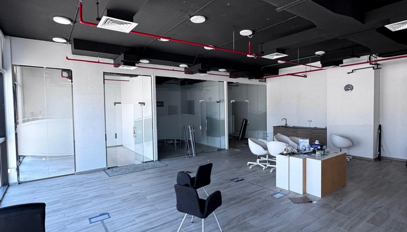 A modern, mostly empty office space with glass-walled rooms, a few chairs, a desk, and scattered office supplies on the tile floor.