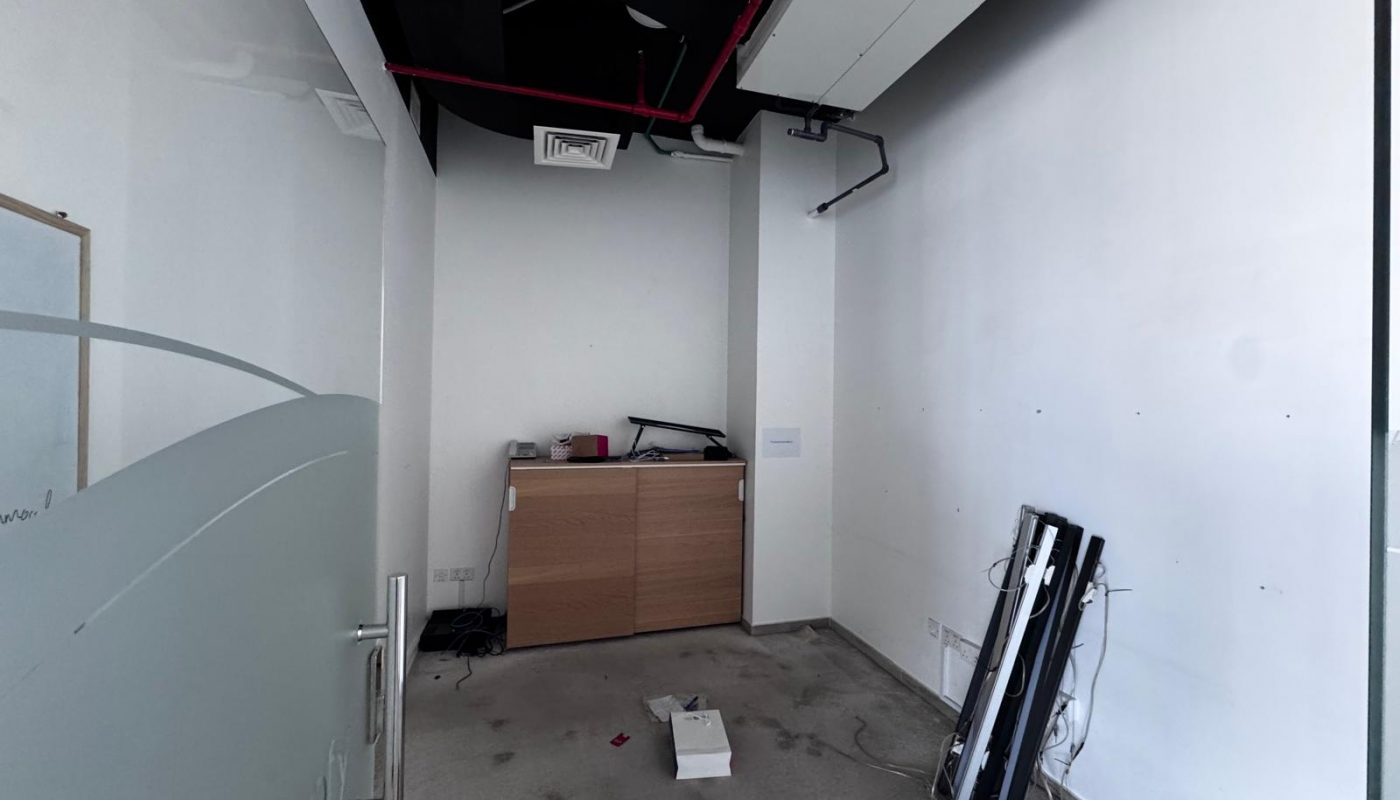 Small, mostly empty office space with a wooden cabinet, some scattered debris on the floor, detached light fixtures, and exposed ceiling ducts.