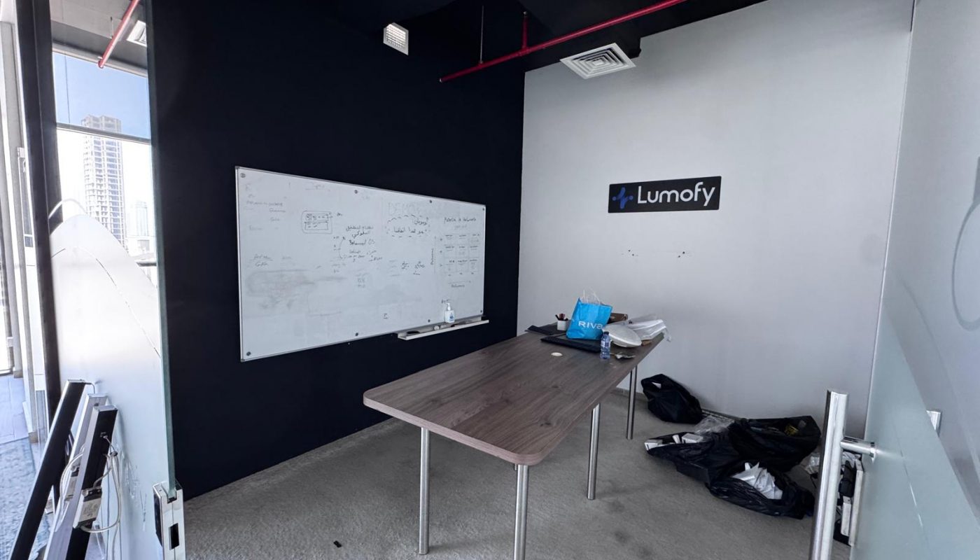 Small office room with a whiteboard covered in notes, a rectangular table with bags and boxes on it, and a "Lumofy" sign on the wall.