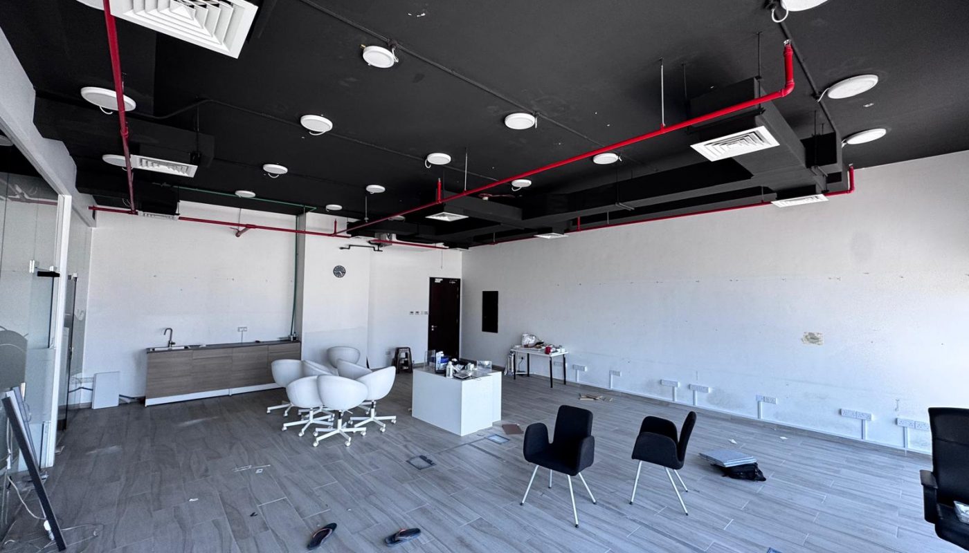 Modern office space with a black ceiling, red pipes, scattered chairs, a small sink area, and various items on a light wood floor. The room appears mostly empty and unfinished.