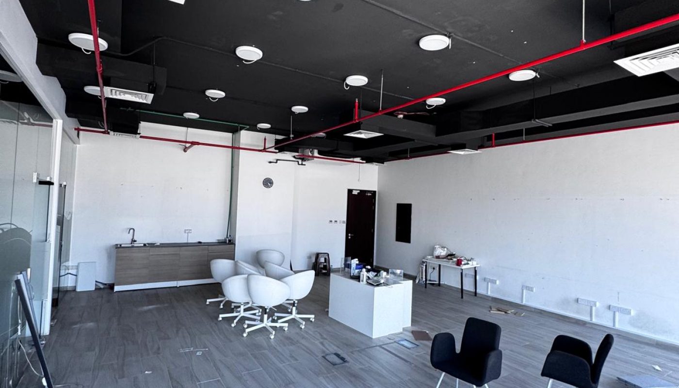 Modern office space with gray wood flooring, scattered chairs, a small white desk, exposed black ceiling with red pipes, and some unpacked items along the walls.