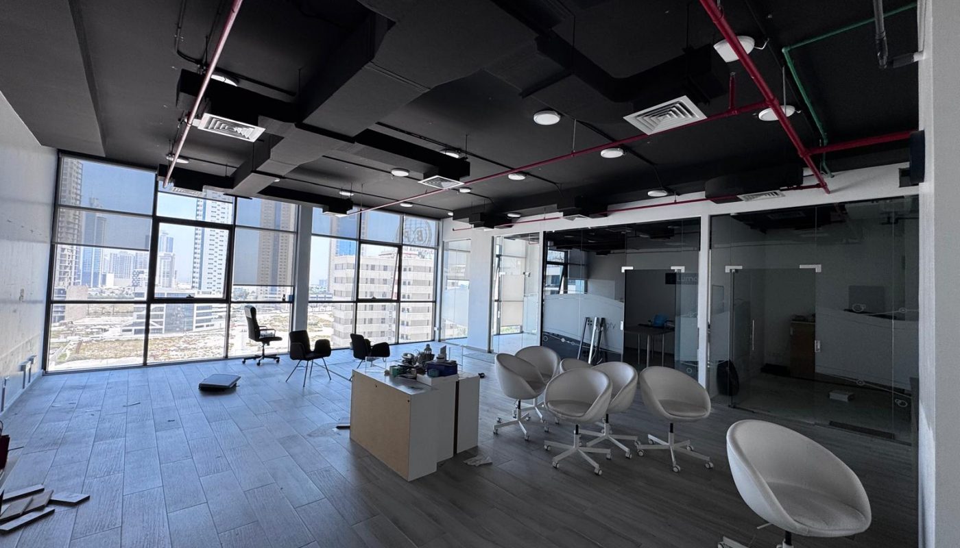 Modern office space with large windows, white chairs, a desk in the center, and scattered office items. The room appears mostly empty and partially disorganized.