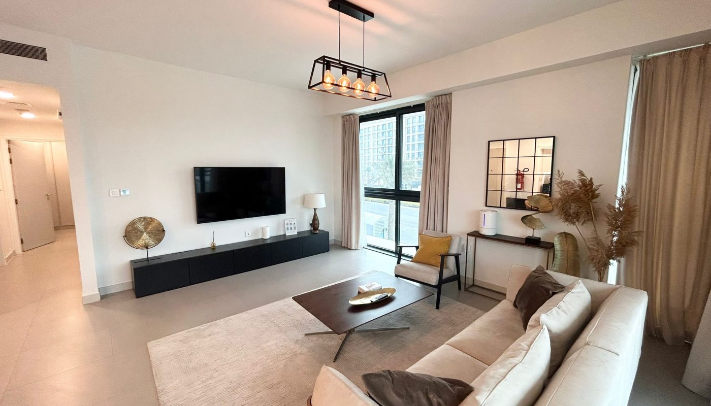Modern living room with beige sofa, coffee table, TV mounted on wall, large window with curtains, neutral decor, and light gray rug on tiled floor.