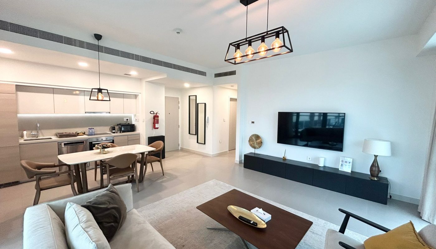Modern open-plan living room and kitchen with neutral tones, a wall-mounted TV, dining table, sofa, and large windows allowing natural light.