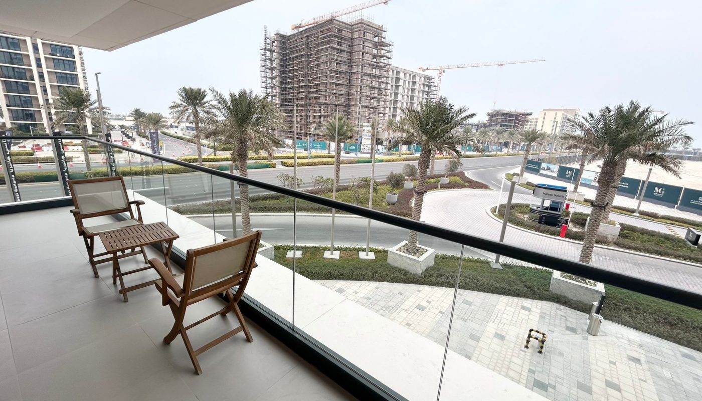 View from a balcony with two wooden chairs, overlooking palm trees, a road, and several buildings under construction.