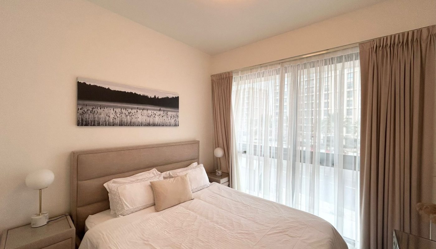 A neatly made bed with white bedding is centered in a bright bedroom with large windows, beige curtains, two bedside tables, and a framed landscape photo above the headboard.