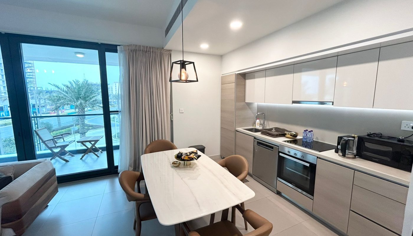 Modern kitchen and dining area with a white table, four chairs, built-in appliances, and a view of a balcony with outdoor seating and palm trees through large glass doors.