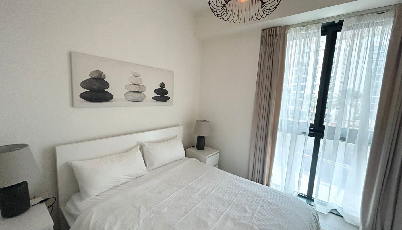 A modern bedroom with a neatly made bed, two bedside tables with lamps, a pebble artwork above the bed, and a large window with sheer curtains letting in natural light.