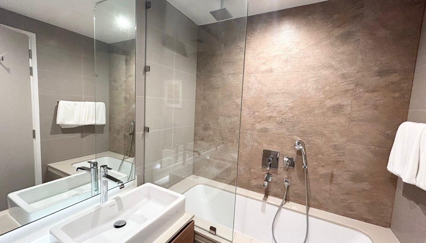 Modern bathroom with a white sink, a glass-enclosed bathtub and shower, wall-mounted faucets, large mirror, and beige tiled walls.