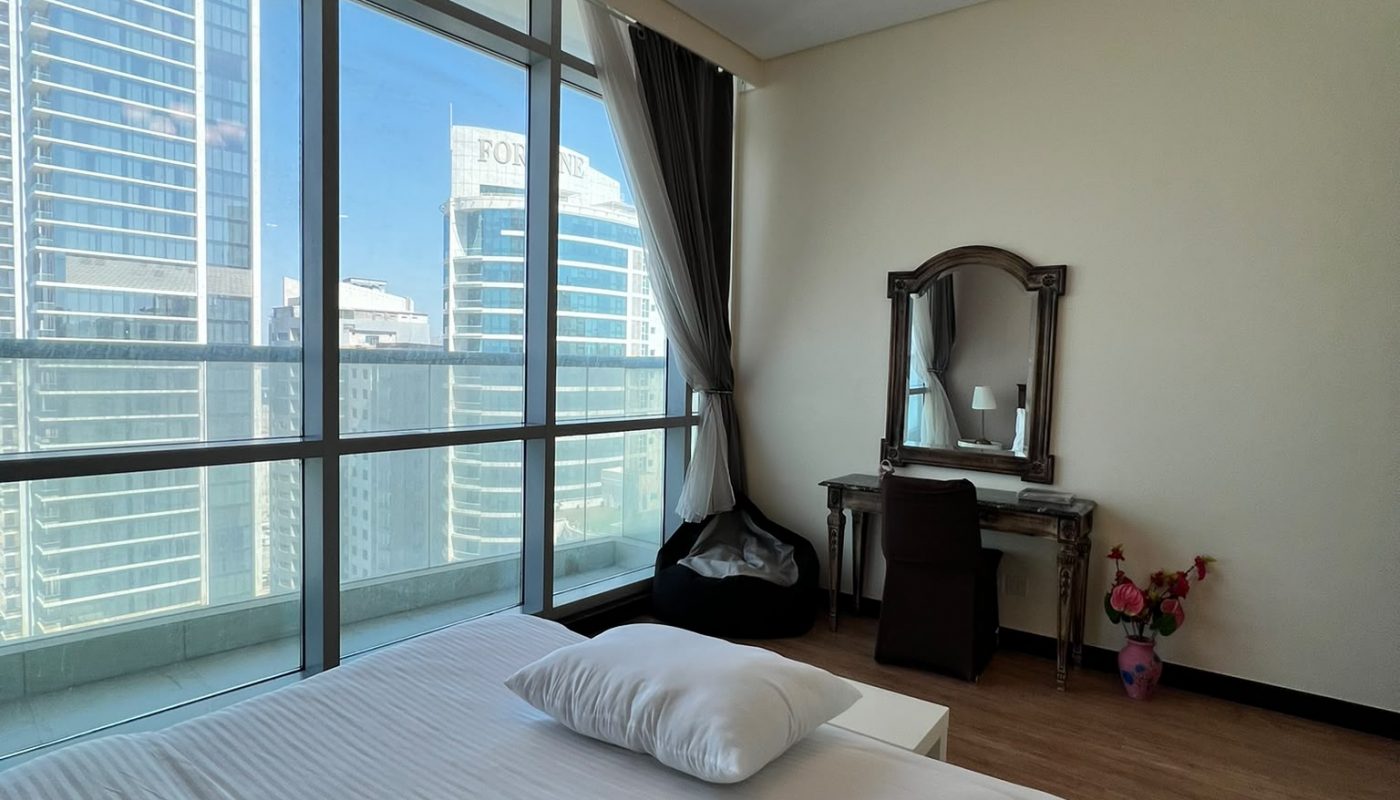 A modern bedroom with a large bed, a vanity table, floor-to-ceiling windows, city views, wooden floor, and a ceiling light fixture.