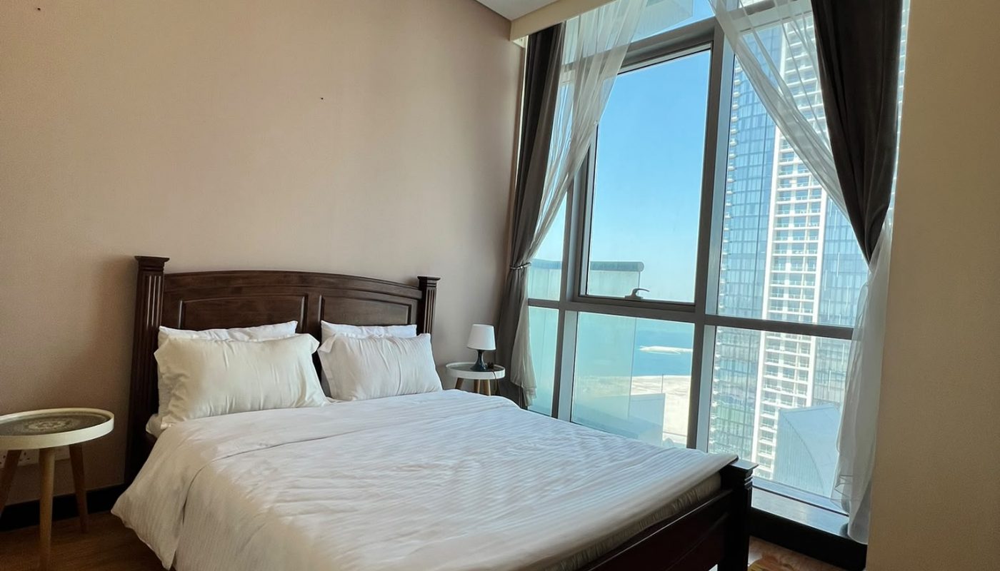 A bedroom with a double bed, white bedding, a chandelier, a side table, lamp, wooden floor, patterned rug, and large windows with city views and curtains.