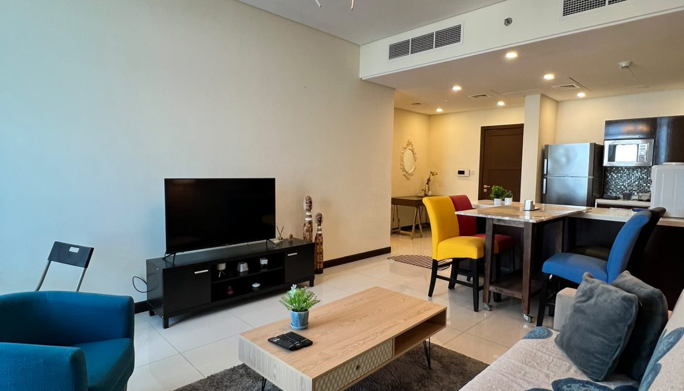 Modern living room with a sofa, coffee table, TV on a stand, blue and yellow chairs, and an open kitchen area in the background under a ceiling light fixture.