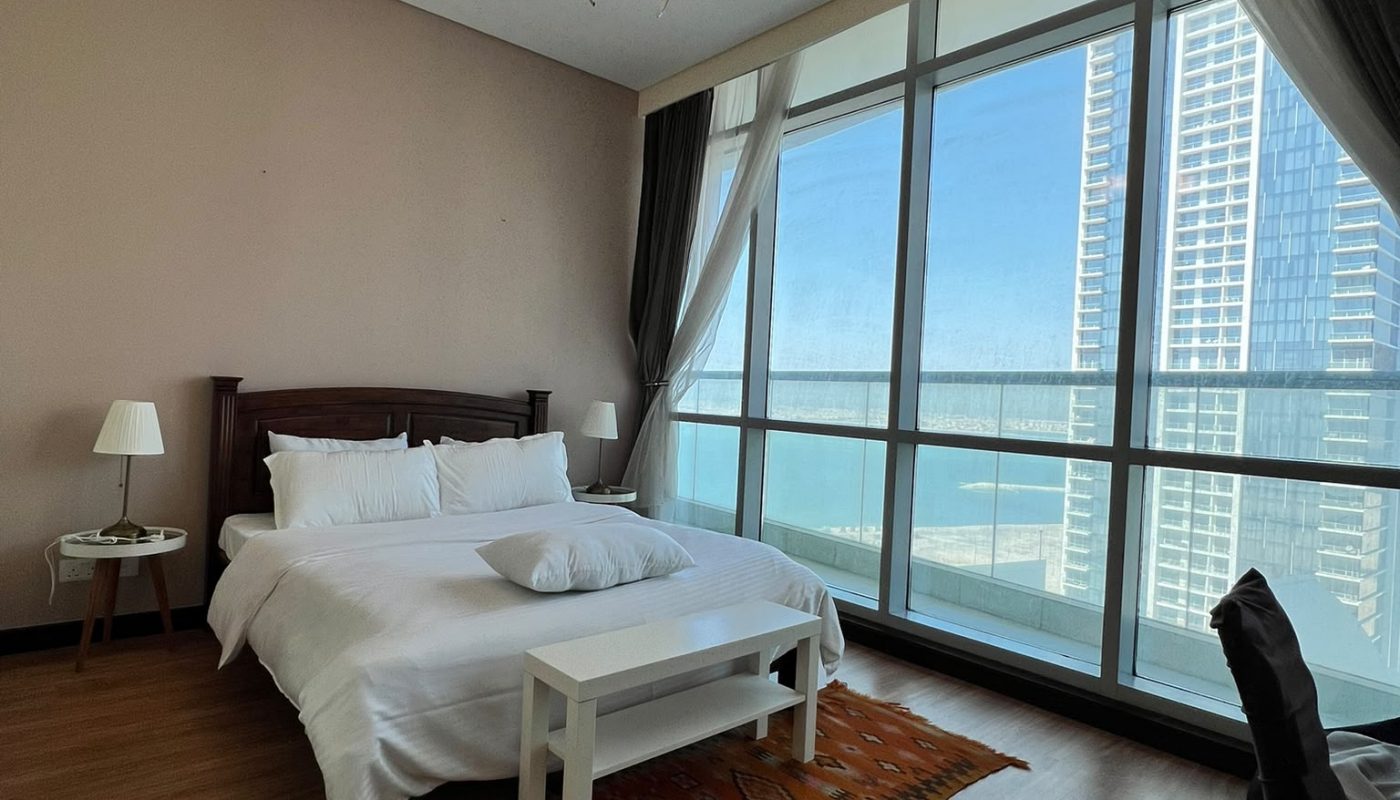 A modern bedroom with a double bed, bedside tables, a patterned rug, and large floor-to-ceiling windows offering a city and ocean view.