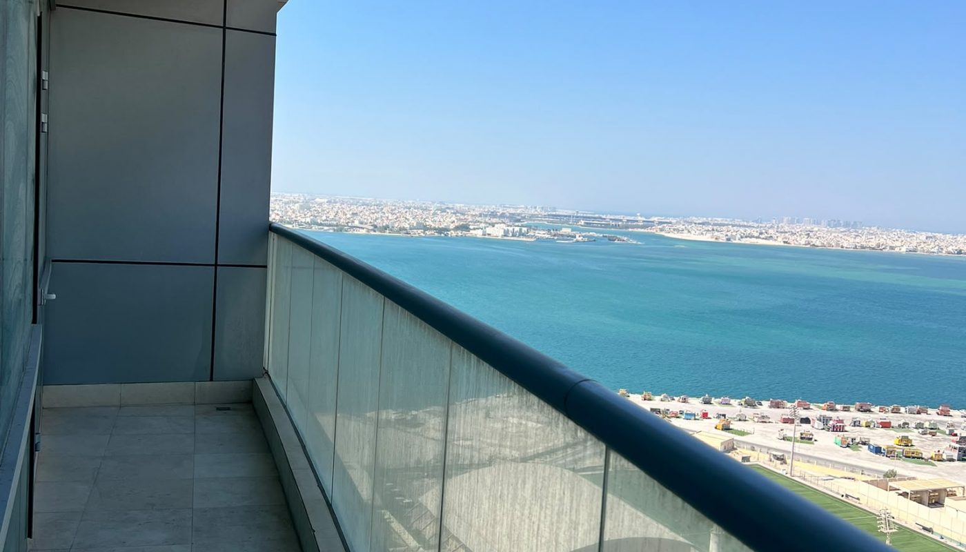 A high-rise balcony with glass railing overlooks a cityscape, a beach, and a blue sea under a clear sky.
