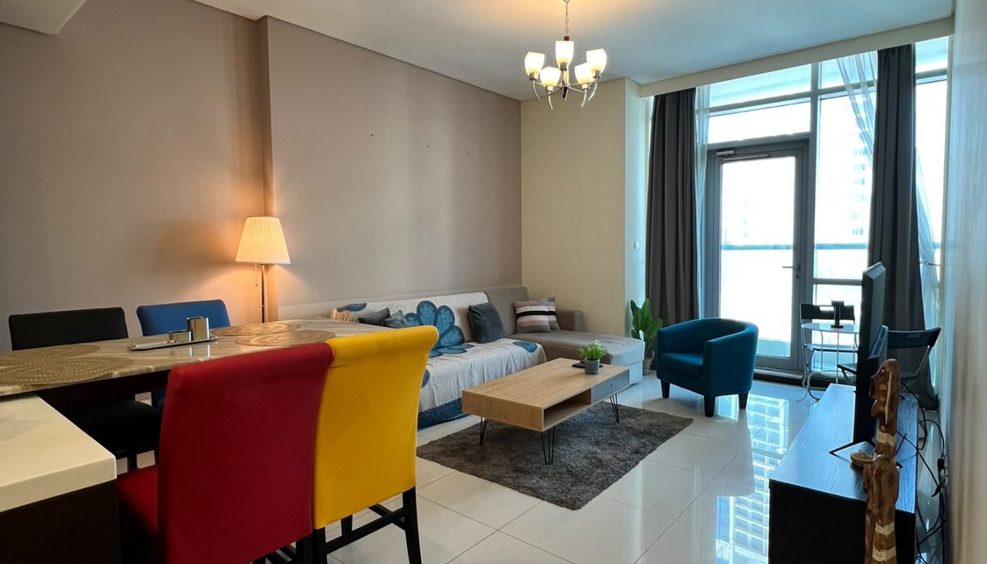 Modern living and dining area with a sofa, colorful chairs, coffee table, lamp, TV, and large window letting in natural light.