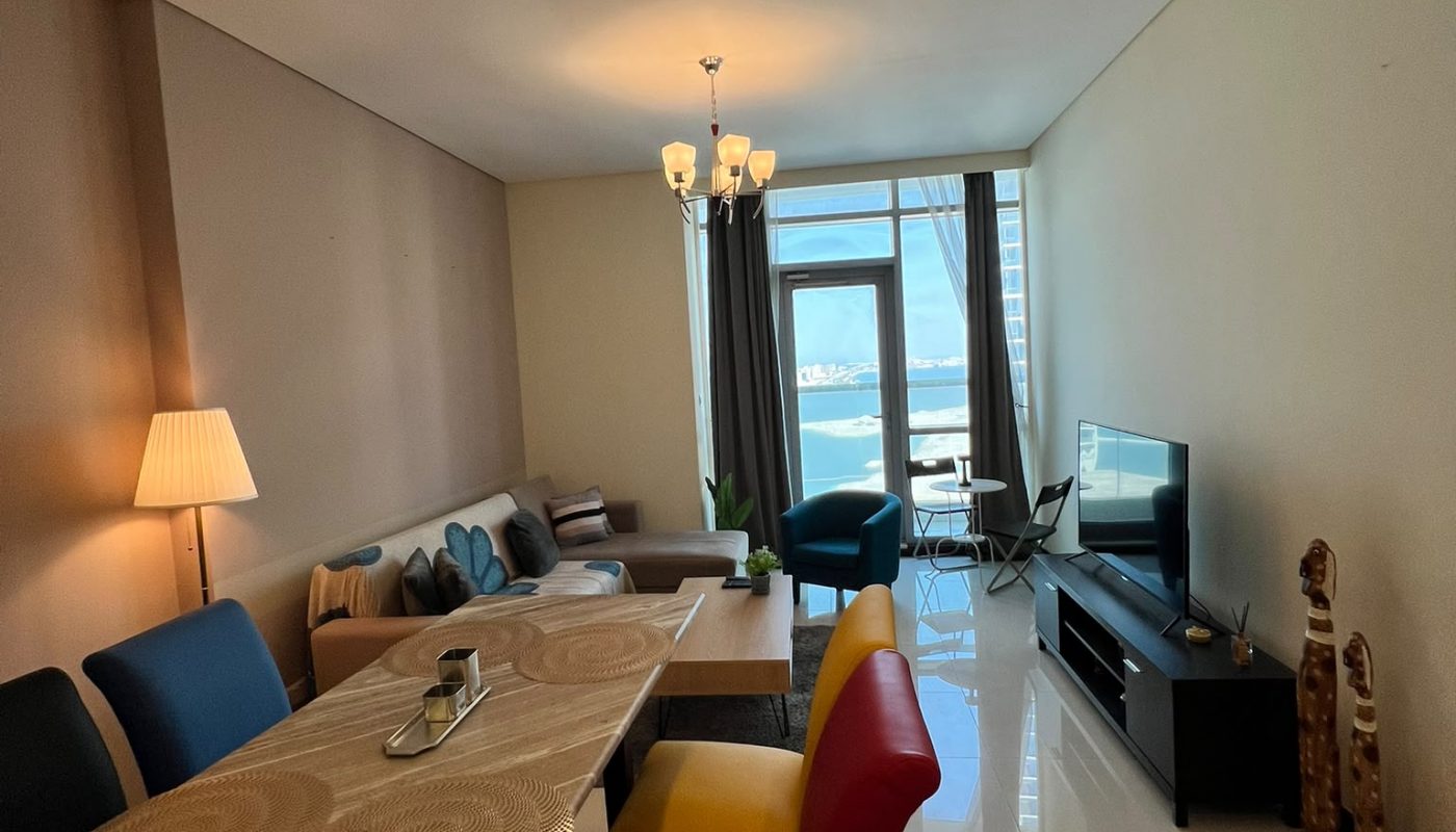 Modern living and dining area with a beige sofa, colorful chairs, TV, decorative items, and large window with curtains letting in natural light.