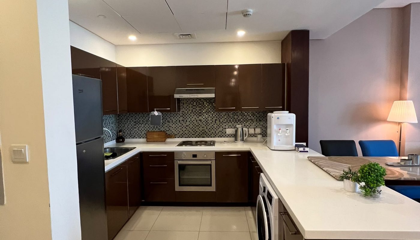 Modern kitchen with brown cabinets, stainless steel appliances, washing machine, white countertop, and a dining area with blue chairs in the background.