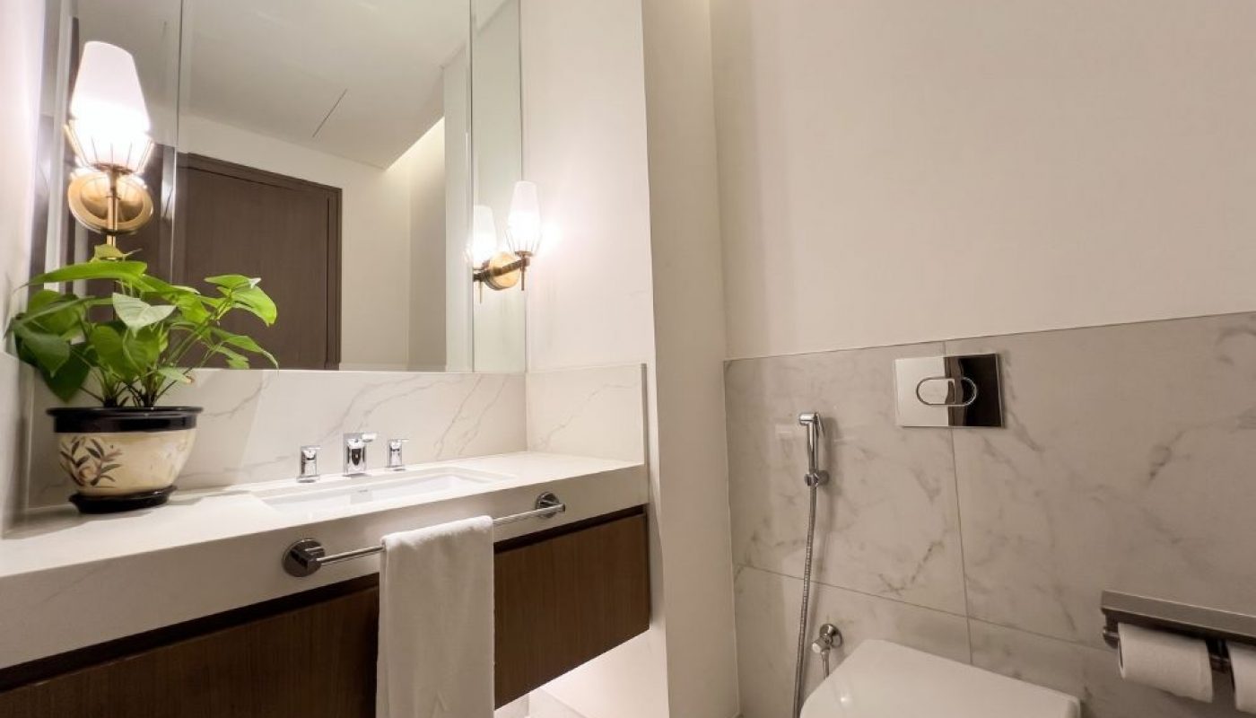 Modern bathroom with a wall-mounted sink, mirror, potted plant, toilet, bidet sprayer, and towel hanging on the vanity. Marble tile walls and soft lighting.
