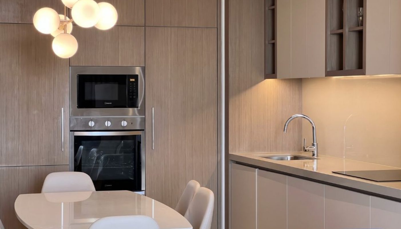 Modern kitchen with light wood cabinets, built-in oven and microwave, a dining table with chairs, round pendant light, and a sink on the right.