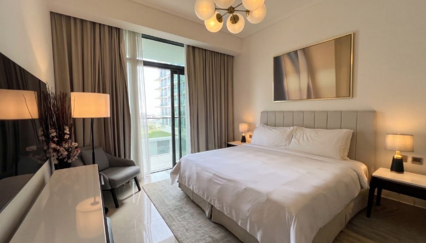 Modern bedroom with a large bed, white linens, two bedside tables with lamps, a chair near a window, and a balcony view with buildings visible outside.