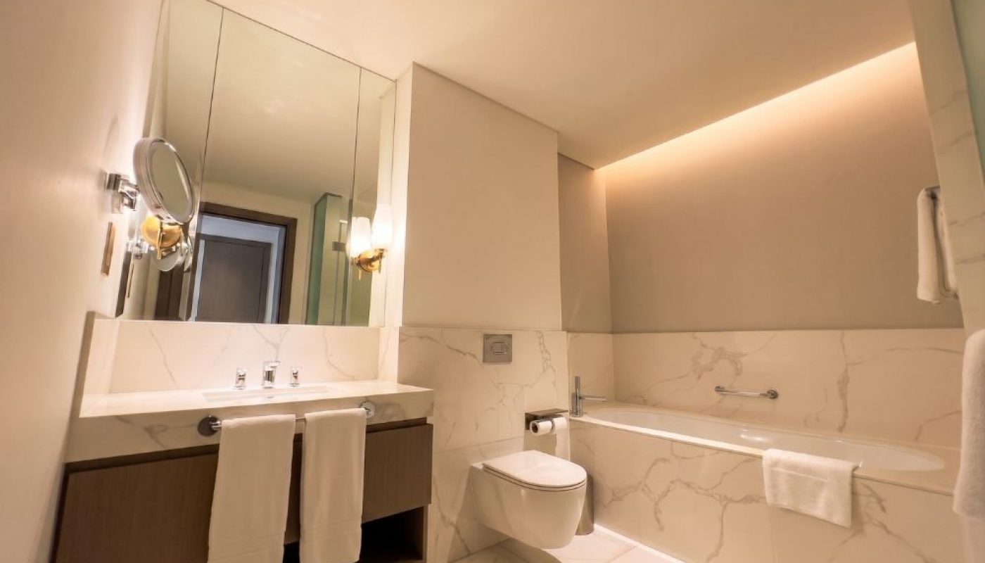 Modern bathroom with a double sink, wall mirror, mounted lights, wall-hung toilet, bathtub, and marble surfaces; towels are neatly hung on racks.