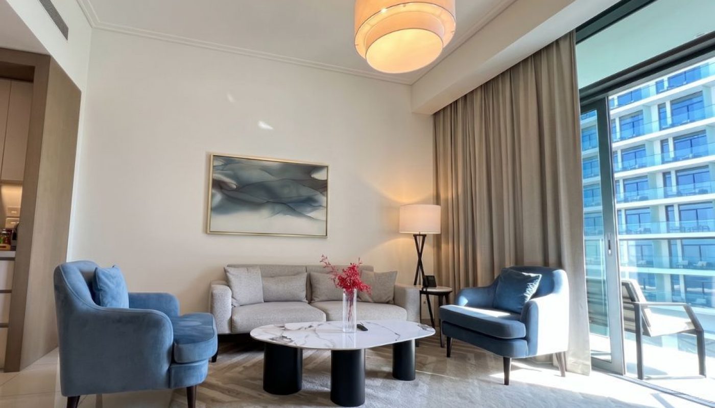 A modern living room with a gray sofa, two blue armchairs, a round marble coffee table, and a large window opening to a balcony with city views.