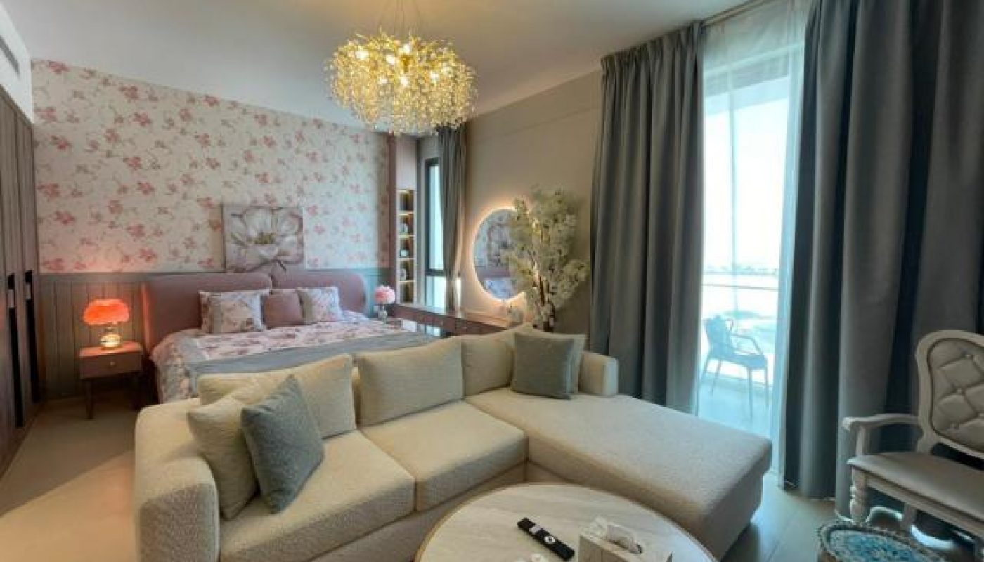 A modern bedroom with a floral accent wall, a large bed, a cream sectional sofa, a round coffee table, and floor-to-ceiling windows with gray curtains overlooking a balcony.